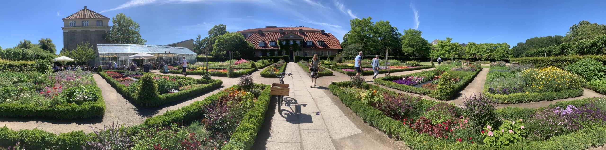 Botanical_Garden_Denmark Πανόραμα του Βοτανικού Κήπου στη Δανία με πολύχρωμα λουλούδια, οργανωμένα μονοπάτια και ένα ιστορικό κτίριο στο βάθος